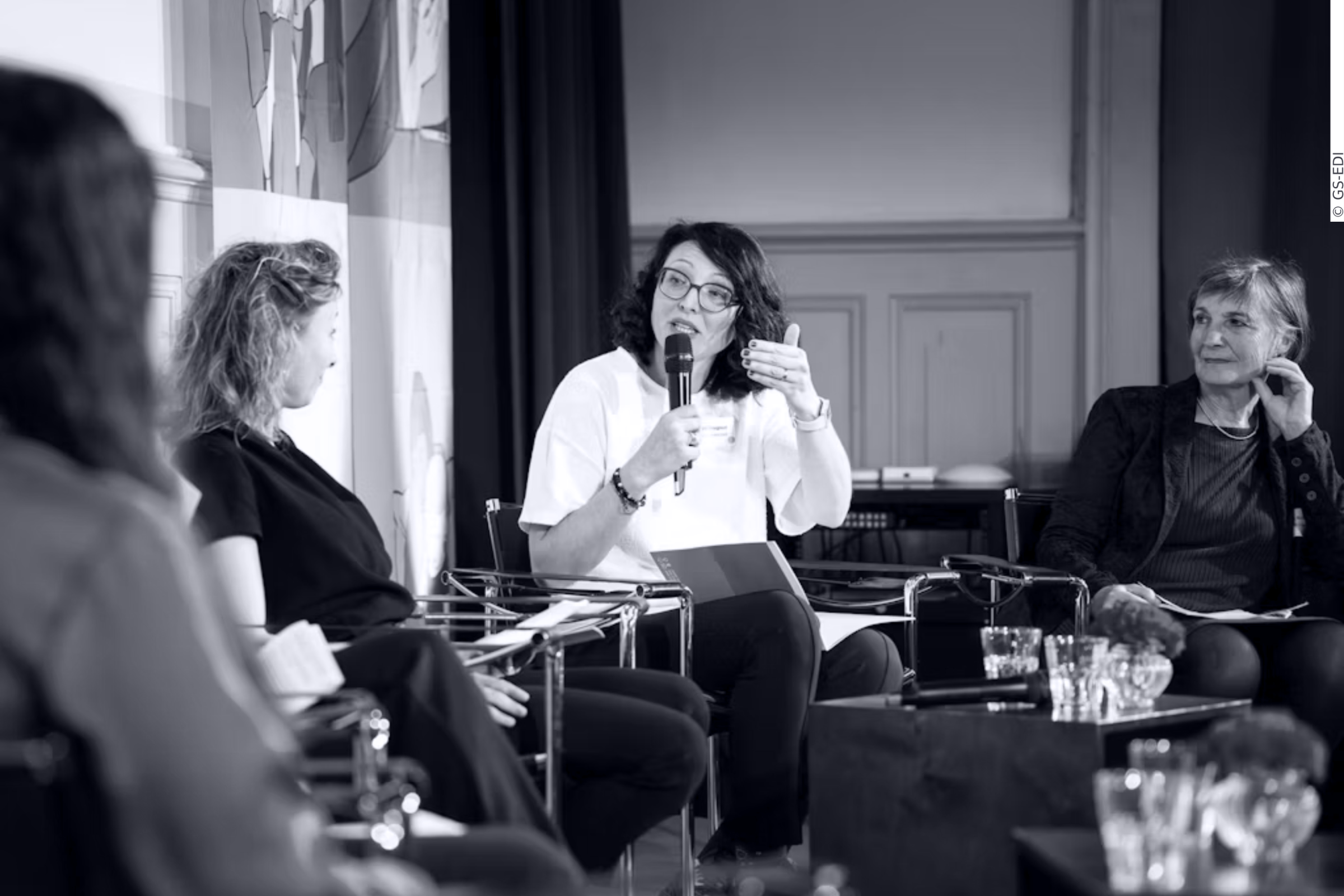 three women are sitting on stage, two have longer and curly hair, one has short and straight hair, the woman in the middle is speaking into a microphone, the colors are black, white and grey